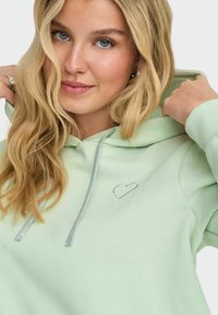 Felpa verde con una texture morbida, presenta un piccolo cuore ricamato in argento e stringhe argento sul cappuccio.