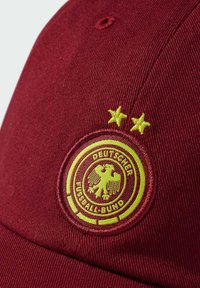 Crvena kapa izrađena od teksturiranog materijala koja sadrži okruglu žutu izvezenu oznaku s orlom, natpis "DEUTSCHER FUSSBALL-BUND" i dvije zvijezde.