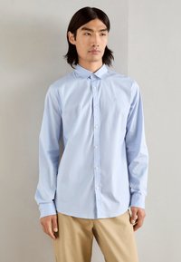 Chemise bleu clair à manches longues, en cotton, avec un petit logo sur la poitrine, associée à un pantalon beige.