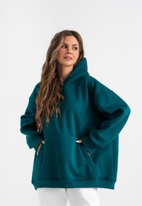 Felpa oversize di colore teal in tessuto morbido, con tasca frontale e cappuccio regolabile con coulisse. Texture liscia con polsini a costine.