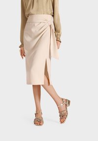 Gonna avvolgente beige con spacco laterale e dettaglio con nodo; abbinata a sandali metallici con lacci e una blusa di seta oliva.