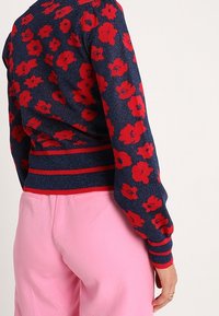 Marinblå tröja med röd blommönster och randig midja, kombinerad med högmidjade ljusrosa byxor på en stående person.