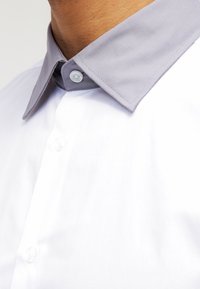 Camicia bianca con colletto grigio. Il colletto presenta un tessuto a contrasto e un bottone. Texture liscia, design classico con bottoni e dettagli ben definiti.