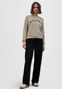 Beżowy sweter dziany z haftowanym na czarno napisem "MILANO"; w połączeniu z czarnymi spodniami o szerokich nogawkach i białymi tenisówkami. Casualowy strój o luźnym kroju.