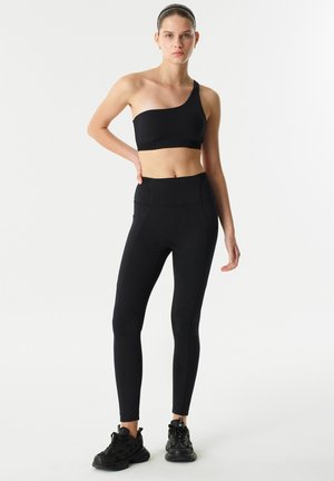 Černý sportovní outfit obsahující crop top na jedno rameno a přiléhavé legíny, vyrobený z elastického materiálu s hladkým povrchem.