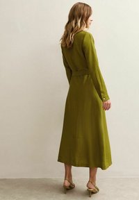 Lång, olivgrön klänning i lätt tyg, med långa ärmar, midjeband och kjollängd nedanför knät. Enkla manchetter.