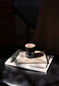 Svart glasljus med trälock, prytt med vita geometriska mönster. Positionerat på en beige duk ovanpå staplade tidningar på en mörk yta.