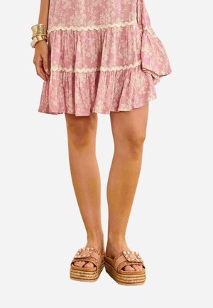 Mujer con un vestido rosa con volantes florales y sandalias tipo slide con plataforma en color beige con hebilla decorativa y suelas trenzadas.