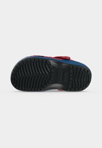 Semelle noire à crampons d'une chaussure Crocs rouge et bleue vue par en dessous sur fond blanc.