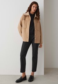 Veste chemise en mélange de laine beige avec col, poches avant et fermeture à boutons ; associée à un pantalon slim noir et des mocassins noirs.