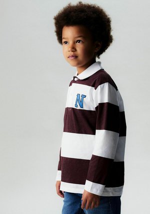 Langermet polostoff med burgunder- og hvite striper og en blå bokstav "N" på brystet, kombinert med blå denimjeans.