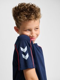 Młody chłopiec z kręconymi włosami, ubrany w granatową koszulkę z czerwonym wykończeniem i dwoma białymi naszywkami na rękawie, nieśmiało się uśmiecha i patrzy w bok.