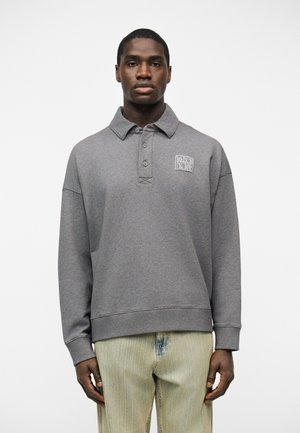 Młody mężczyzna ubrany w szarą bluzę z kołnierzykiem z logo "Maison Kitsuné", stojący na jednolitym białym tle.