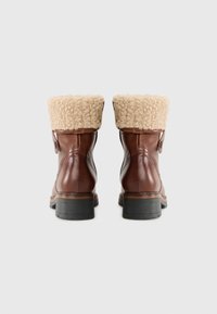 Brązowe skórzane buty z beżowym, puszystym kołnierzem ze shearlingu, okrągłym czubkiem i kontrastującą czarną gumową podeszwą. Posiadają szczegóły w postaci bocznych guzików.