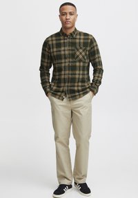 Camisa de franela de cuadros verdes y amarillos con cuello abotonado, manga larga y bolsillo en el pecho, combinada con pantalones beige y zapatos negros.