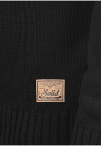 Musta neulepaita, jossa on ribbitekstuuri ja nahkapläkkijossa lukee "Solid United Traditions 1993" alaosassa.