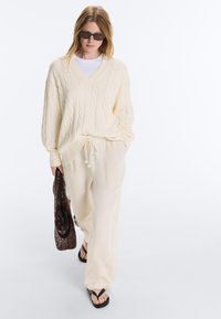 Femme marchant en pull crème à torsades, pantalon large assorti, tongs noires, lunettes de soleil foncées, tenant un sac cabas marron à motifs.