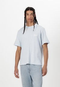Light blue cotton t-shirt with short sleeves, round neck, and small white text logo on the chest. Paired with light denim jeans.