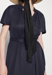 Mujer que lleva un vestido envuelto azul marino con mangas cortas y un cinturón de tela a juego, bufanda negra atada al cuello y sombrero de paja.