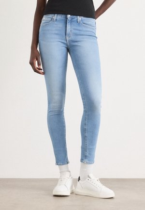 Persoon die lichtblauwe skinny jeans, witte sokken en witte sneakers draagt, staand op een neutrale vloer tegen een effen achtergrond.