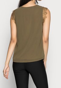 Oliivinvihreä hihaton paita, jossa on pitsikoristeet olkapäillä, pehmeä kangas, pyöreä kaula-aukko ja kaartuva helma, yhdistetty mustiin legginsseihin.