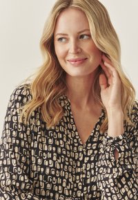 Juoda ir smėlio spalvos raštuota bliuskutė su V formos kaklu ir surinktomis rankovėmis. Medžiaga yra lygi, su abstrakčių formų kartojančiu raštu.