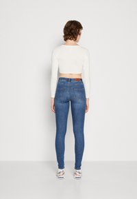 Kék farmer skinny nadrág magas derékkal, hátsó zsebekkel és márkázott bőrpólóval. Fehér, rövid fazonú, hosszú ujjú toppal párosítva.