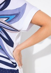Top de manga corta con un fondo blanco que presenta patrones abstractos en tonos azules, morados y beige, con una textura suave y detalles laterales.