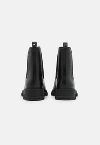 Mustat nahkaiset chelsea-kengät, joissa on sileä pinta, paksu kumipohja ja bränditunnus nauhoissa. Kengissä on kohokuvioitu logo kannassa.