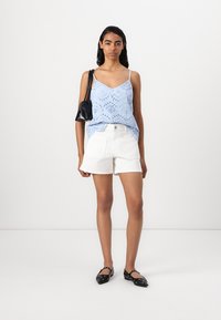 Top de algodão azul claro com detalhes de ilhós, combinado com calções de algodão brancos. A modelo usa sapatilhas pretas e carrega uma pequena mala preta.