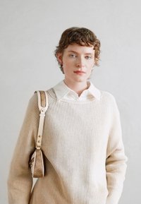 Suéter de canalé beige con escote redondeado, combinado con una camisa blanca de cuello. Un pequeño bolso con un acento estampado se lleva al hombro.