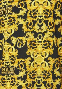 Tissu noir avec des motifs complexes de volutes baroques dorées et des motifs floraux, avec le texte "Versace Jeans Couture" en or.