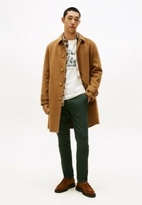 Cappotto di lana marrone chiaro con grandi bottoni, indossato sopra una t-shirt bianca con grafica e pantaloni verdi. Scarpe in suede marroni completano l'outfit.