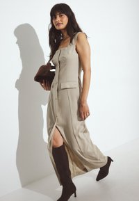 Beige linned maxi kjole med knapfront, firkantet halsudskæring og sideslids; parret med brune knæhøje støvler og en matchende clutch.