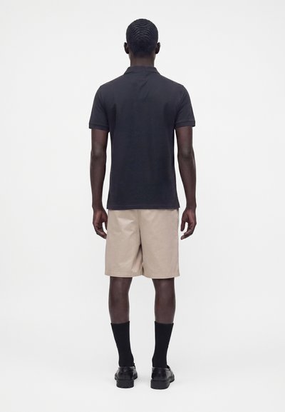 Homme debout de dos, portant un polo noir à manches courtes, un short beige, des chaussettes noires et des chaussures noires sur un fond blanc uni.