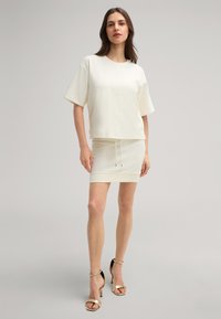 Damenoberteil in hellem Beige mit kurzen Ärmeln, kombiniert mit einem passenden, figurbetonten Rock, der über einen Kordelzug und eine glatte Textur verfügt. Goldene Sandalen mit Absatz.