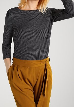 Persoon die een zwart-wit gestreept shirt met lange mouwen draagt, ingestopt in een hooggetailleerde okergele broek met een gestrikte riem.