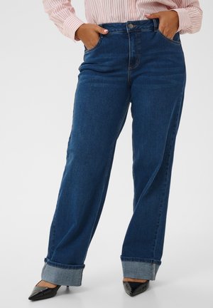 Personne portant un jean large bleu marine à revers, une chemise rayée rose clair et des escarpins noirs à bouts pointus, les mains dans les poches.