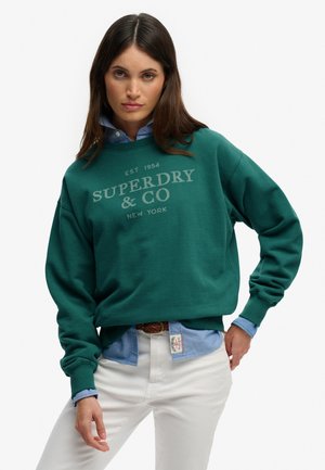 Femme portant un sweat-shirt vert Superdry & Co par-dessus une chemise bleue boutonnée, avec un pantalon blanc, posant avec une main sur la hanche devant un fond blanc.