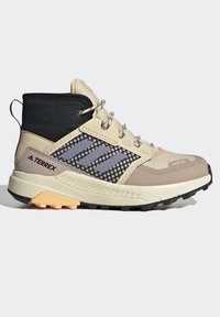 Zapato de senderismo de media altura en color beige y negro, con paneles de malla, superposición sintética y suela de goma. Presenta tres rayas grises y acentos texturizados.