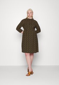 Vestido preto com bolinhas amarelas, mangas longas, decote franzido e comprimento até ao joelho. Usado com sandálias de salto castanhas.