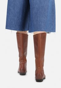 Botas de cuero marrón hasta la rodilla combinadas con unos jeans de mezclilla azul recortados con dobladillos deshilachados, vistas desde atrás sobre un fondo blanco.