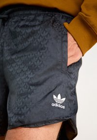 Svarta shorts med ett mönstrat mönster, med en vit Adidas-logotyp vid kanten. Sidoficka synlig, dragsko i midjan.