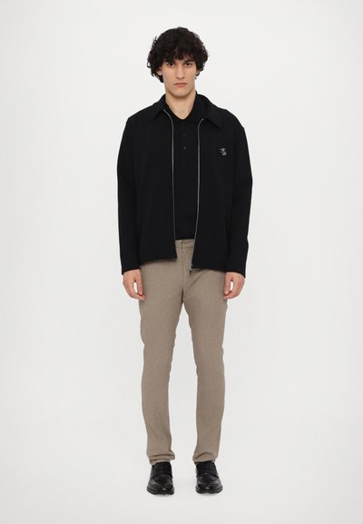 Svart zip-up jacka med krage, bärs över en svart polo skjorta. Beiga åtsittande byxor. Modellen har på sig svarta skor och står på en vit bakgrund.
