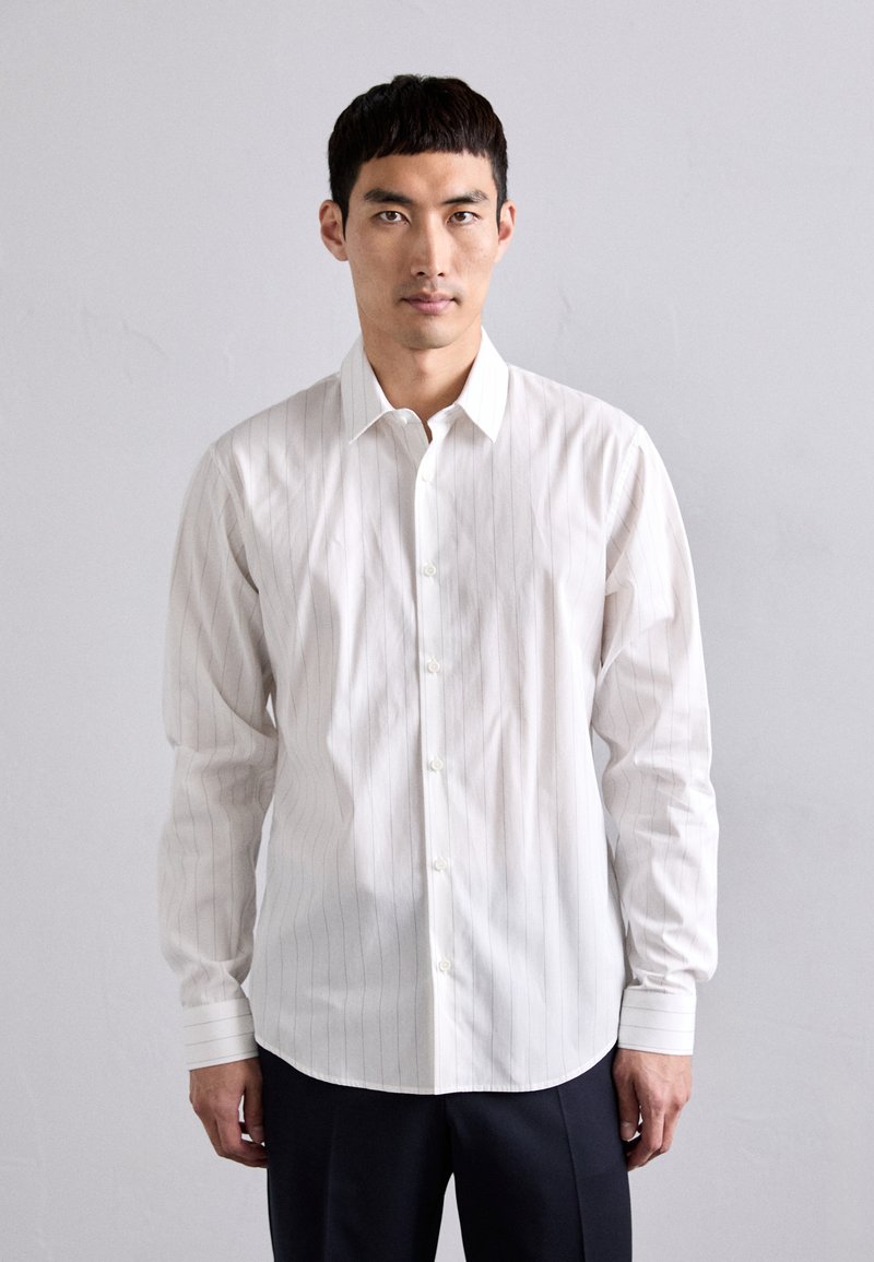 Camisa blanca de botones con sutiles rayas verticales, mangas largas y un cuello clásico. Puños ajustables y textura de algodón suave.