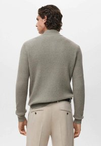 Maglione a coste di un beige chiaro con un colletto alto, abbinato a pantaloni beige su misura con tasche posteriori e dettagli sottili.