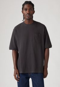 Tmavě šedé oversized tričko s krátkými rukávy, kulatým výstřihem a jedinou náprsní kapsou; vyrobené z hladkého materiálu s minimalistickým designem.