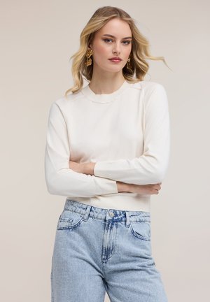 Lyshåret kvinde iført guldringe, hvid langærmet top og lyseblå højtaljede jeans med rhinsten detaljer, armene krydset.