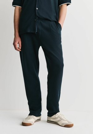 Mann trägt schwarze strukturierte Hose, ein passendes kurzärmeliges Hemd mit Knöpfen und weiße Sneakers mit braunen Zehenschutzkappen, steht mit einer Hand in der Tasche.