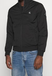 Giacca bomber nera con zip, dal tocco morbido, colletto alto, tasche laterali e piccolo logo ricamato sul petto e sulle maniche.